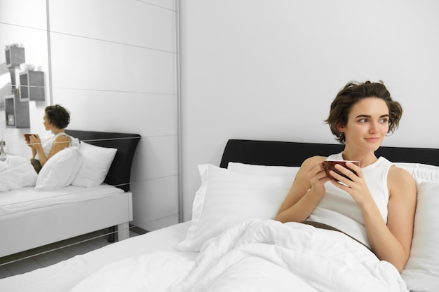 Портрет красивой молодой женщины, отдыхающей в своей спальне, лежащей в постели и пьющей кофе.