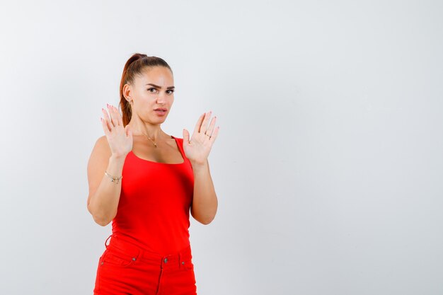 Portrait of beautiful young woman showing surrender gesture in red tank top, pants and looking scared front view