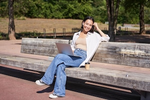 Portrait of beautiful young woman sitting on bench in park using laptop and drinking coffee relaxing