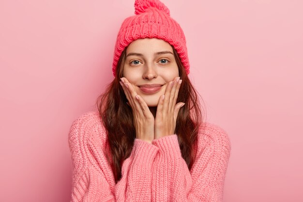 美しい若い女性の肖像画は心地よく微笑んで、両手のひらを頬に保ち、カメラを喜んで見、リラックスした表情をしており、ニットの冬用帽子とセーターを着て、ピンクのスタジオの壁に屋内でモデルを描いています