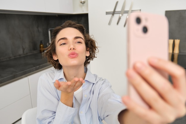 Portrait of beautiful young woman taking selfie on mobile phone at home poses for photo with smartph