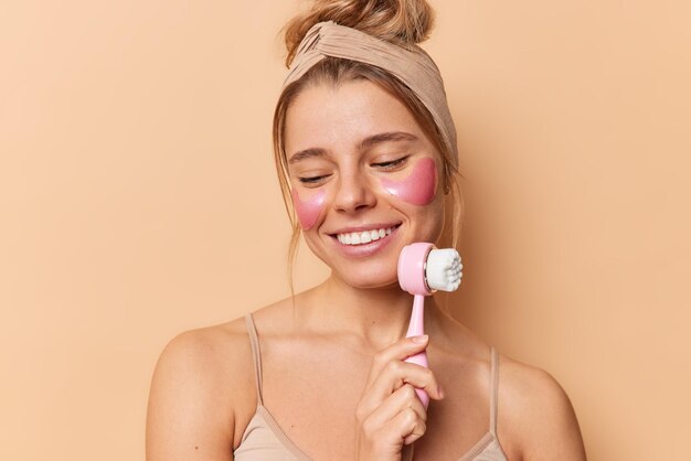 Portrait of beautiful young woman uses face massager takes care of facial skin applies pink hydrogel patches under eyes to remove puffiness wears beige headband and casual t shirt poses indoors