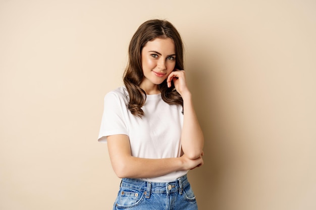 Portrait of beautiful young woman in white t-shirt, smiling and looking happy, posing against beige background.