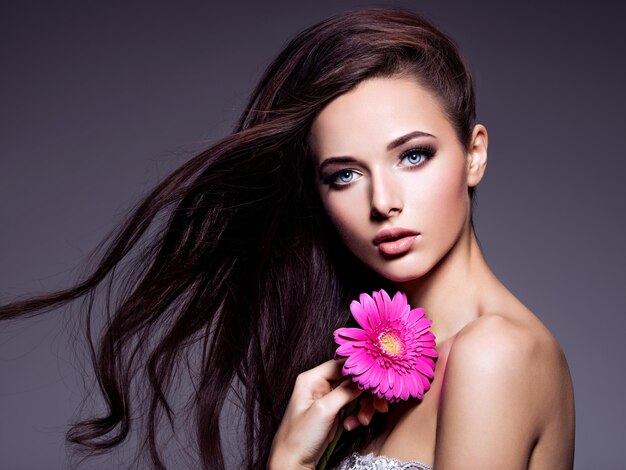 Portrait of the beautiful  young woman with long brown  hair with pink flower posing  over dark wall