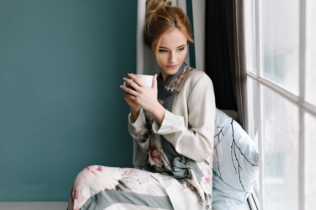 Portrait of beautiful young woman with sensual look through window, sitting on window sill with  mug of coffee in her hands. Turquoise wall. Dressed in silk pajamas with  flowers.