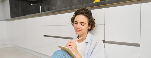 Free photo portrait of beautiful young woman writing in journal adding notes in planner sitting on floor and