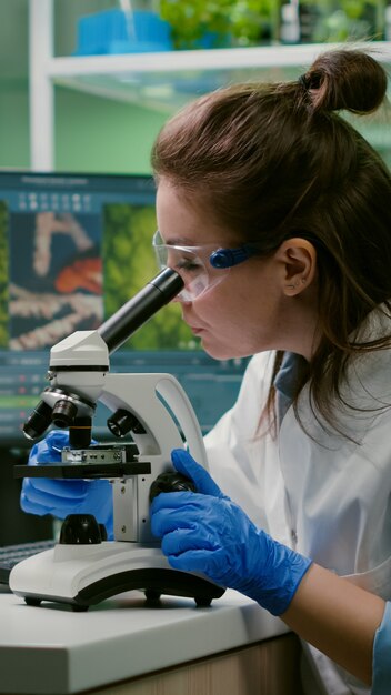 Портрет ученого-биолога в белом халате, работающего в лаборатории экспертизы, смотрящего в микроскоп, анализирующего органический лист ГМО