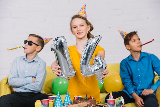 Free photo portrait of a birthday girl showing numeral 14 foil balloon with her two friends blowing party horn