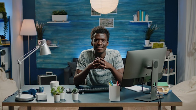 コンピューターとノートブックを持って机に座っている黒人の大人の肖像画。オンラインリモートビジネスの仕事のためのガジェットを持っている間、カメラを見て、笑っているアフリカの民族の男。