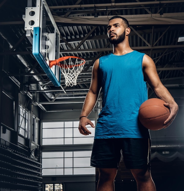 Portrait of Black basketball player holds a ball over a hoop in a basketball hall.