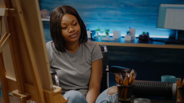 Portrait of black paralyzed artist drawing on canvas while sitting in wheelchair at studio. Woman of african american ethnicity with disability working on modern masterpiece creation