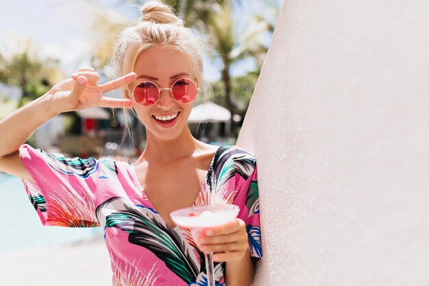 Portrait of blissful blonde woman posing with peace sign and drinking cocktail Happy tanned caucasian girl in bright dress having fun at summer resort