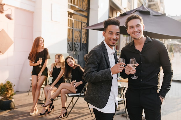 Portrait of blissful women in trendy dresses sits on chairs with handsome men on foreground