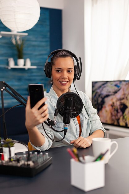 Portrait of blogger woman taking selfie for audience using smartphone working in podcast home studio. Content creator recording new fashion and beauty review and having fun on social media platform.