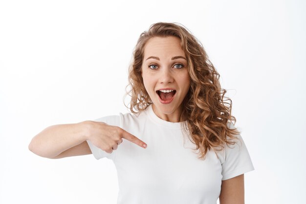 Portrait of blond excited woman, pointing finger at copy space logo, stare with amazed face, showing promo offer impressed, standing over white wall