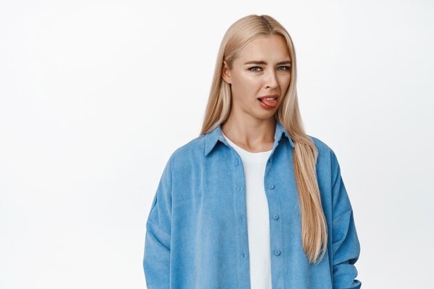 Portrait of blond girl looks disgusted, sticking tongue and grimacing from dislike or bad taste, standing on white