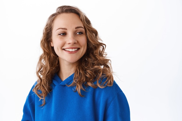 Portrait of blond happy woman look aside at promotional text, smiling satisfied, gazing at something beautiful, white wall