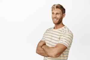 Foto gratuita ritratto di modello maschile biondo con le braccia incrociate sorridente e guardando da parte il prodotto in piedi in maglietta su sfondo bianco