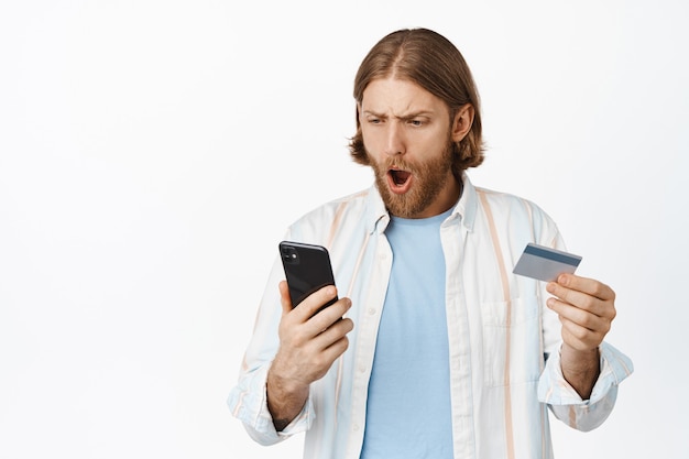 Portrait of blond man looks shocked at his phone screen, bank account, gasping worried, holding credit card, lose money, standing on white