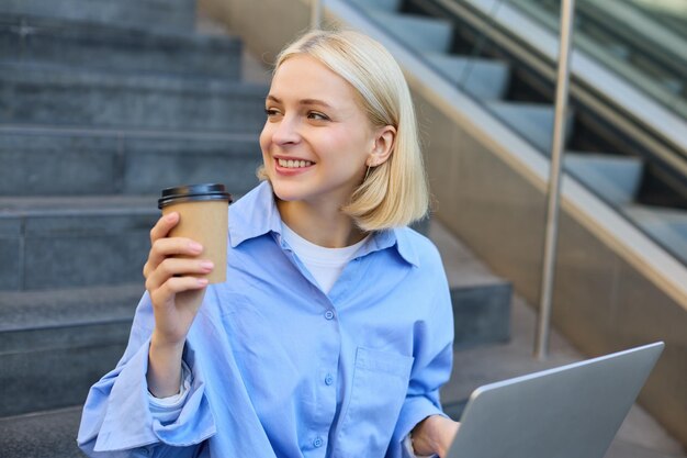 Free photo portrait of blond smiling woman drinking coffee using laptop sitting on campus stairs outside of