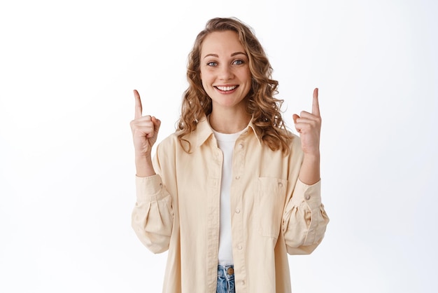Портрет улыбающейся блондинки в стильной одежде, указывающей пальцем на пространство для копирования, демонстрирует рекламный текст, довольный перед камерой, рекомендуя продукт на белом фоне