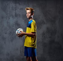 Portrait of blond teenager, soccer player dressed in a yellow uniform holds a ball.