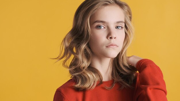 Портрет белокурой волнистой девушки-подростка, выглядящей уверенно изолированной на желтом фоне. Выражение лица