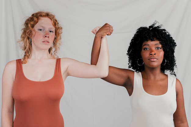 Portrait of blonde and african young women making fist standing against grey backdrop