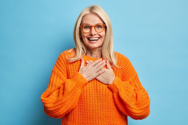 Portrait of blonde female with cheerful expression keeps hands pressed to chest expresses gratitude for heartwarming compliment wears eyeglasses and orange sweater.