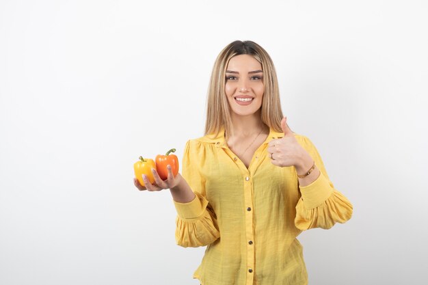 Portrait of blonde girl holding colorful bell peppers and giving thumbs up.