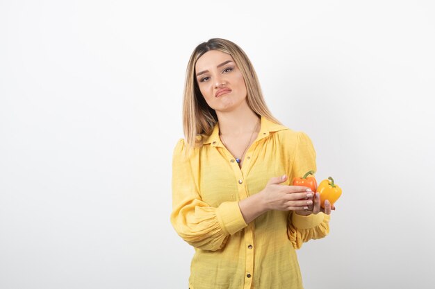 Portrait of blonde girl holding colorful bell peppers on white wall.