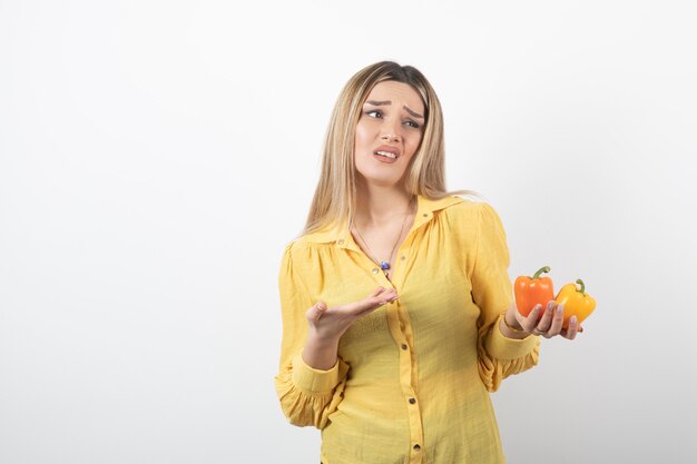 Portrait of blonde girl holding colorful bell peppers on white wall.