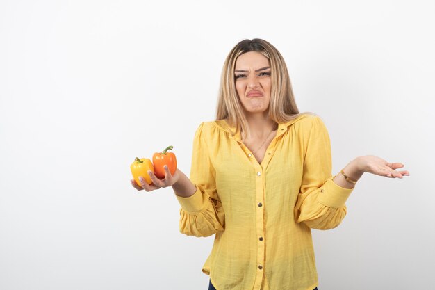 Portrait of blonde girl holding colorful bell peppers with unreadable expression.