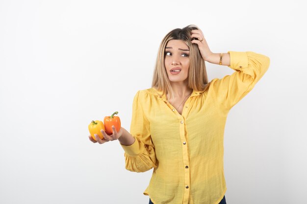 Portrait of blonde girl holding colorful bell peppers with unreadable expression.