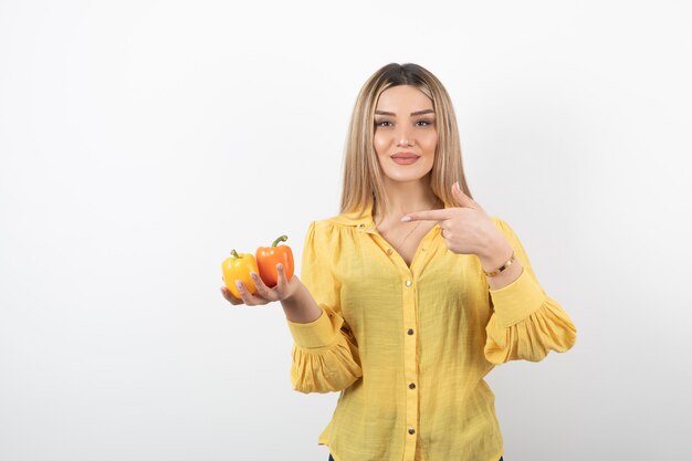 Portrait of blonde girl pointing at colorful bell peppers on white wall.