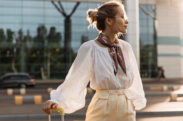 Портрет блондинки в белой блузке, бежевых брюках, коричневом шелковом шарфе и очках, держащей багаж и смотрящей вдаль