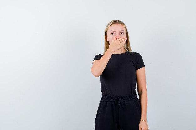 Portrait of blonde lady keeping hand on mouth in black dress and looking scared front view