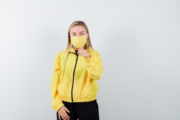 Portrait of blonde lady pointing at camera in tracksuit, mask and looking wondered front view