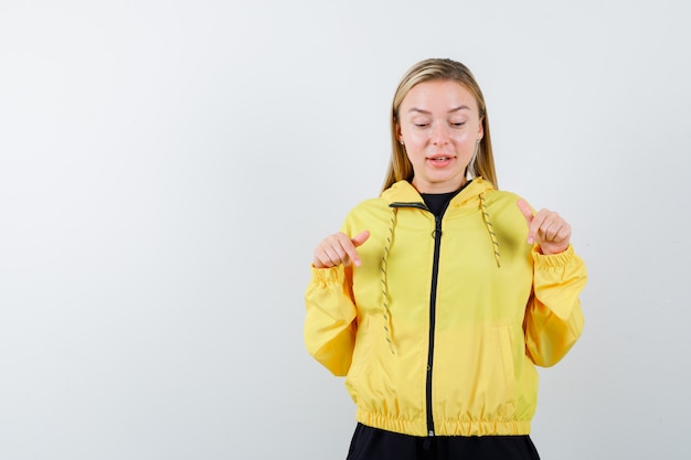 Portrait of blonde lady pointing down in tracksuit and looking astonished front view