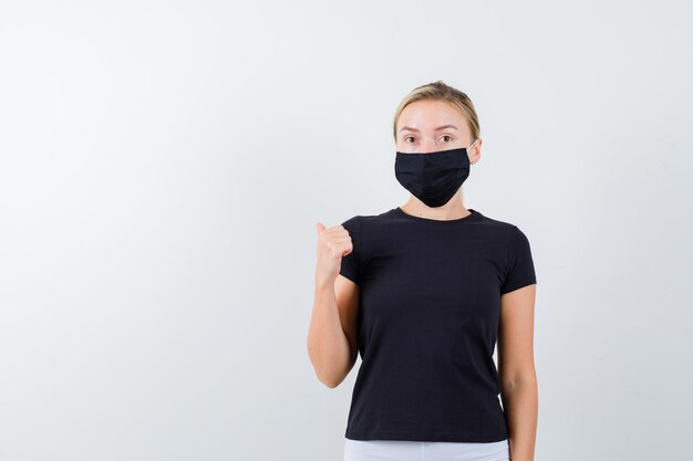 Portrait of blonde lady pointing thumb back in black t-shirt isolated