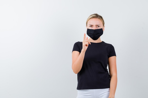 Portrait of blonde lady pointing up in black t-shirt, black mask isolated