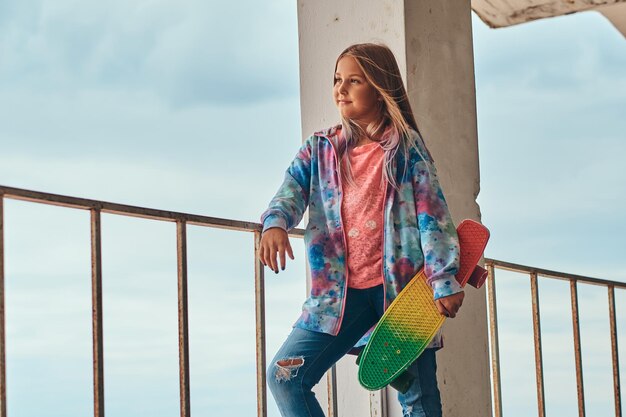 Portrait of a blonde schoolgirl skater posing with a skateboard while leaning on a guardrail against a sky.