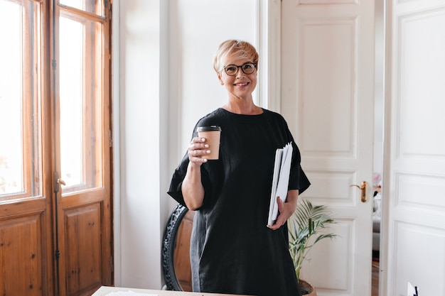 Portrait of blonde woman in eyeglasses and black dress holding tea cup and documents