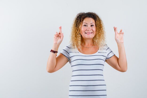 Portrait of blonde woman keeping fingers crossed in striped t-shirt and looking happy front view