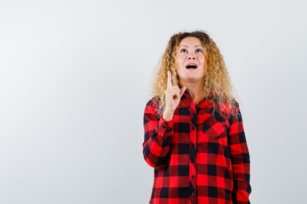 Portrait of blonde woman with curly hair pointing up, looking upward in checked shirt and looking grateful front view