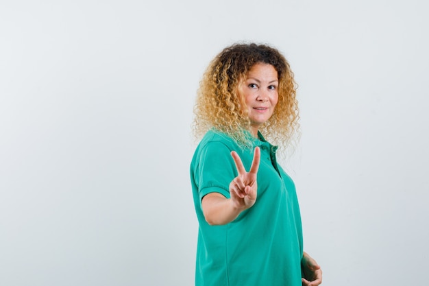 Portrait of blonde woman with curly hair showing peace gesture in green T-shirt and looking confident front view