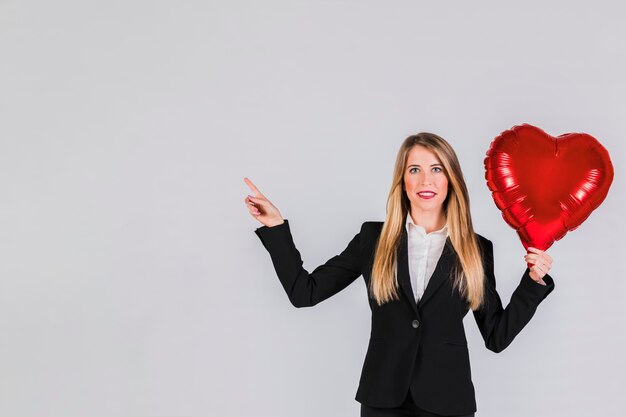 Portrait of a blonde young businesswoman holding red foil balloon in hand pointing her finger