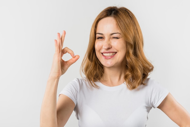 Foto gratuita ritratto di una giovane donna bionda che fa segno giusto che sbatte le palpebre contro il fondo bianco