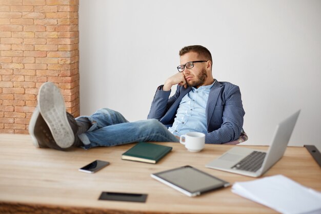 メガネと青いスーツの退屈な大人の白人のひげを剃っていない男性会社のマネージャーの肖像画は、オフィスでの長い一日の後に疲れて、不幸な表情でテーブルに足で座っています。