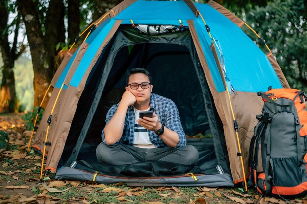Portrait of Bored Asian traveler man glasses using smartphone in tent camping Outdoor traveling camping and lifestyle concept